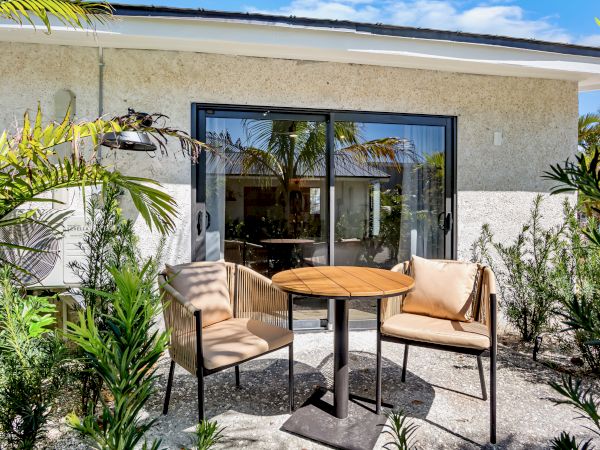 A small outdoor seating area with a round wooden table and beige cushioned chairs sits on a gravel patio, framed by tropical plants and a sliding glass door.
