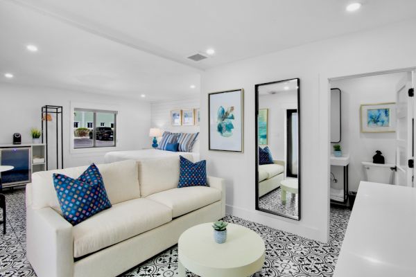 A bright living room with a white sofa, blue pillows, patterned floor, and artwork; a mirror and a small bathroom in view.