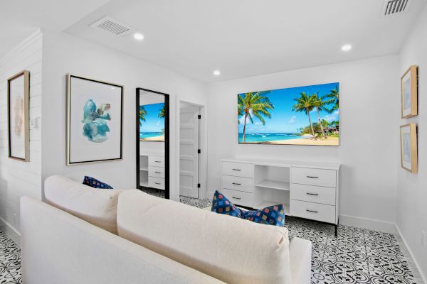 A modern, minimalist room with a couch, tropical beach art on the wall, a door, and patterned flooring, featuring a TV screen showing a beach.