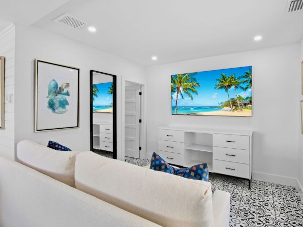 A modern, minimalist room with a couch, tropical beach art on the wall, a door, and patterned flooring, featuring a TV screen showing a beach.