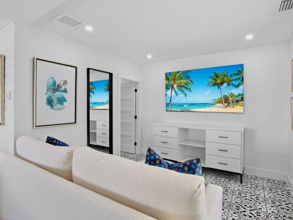 A bright living room with a white sofa, tropical wall art, a flat-screen beach scene, mirrored closet, and patterned black-and-white floor, inviting coastal vibes.