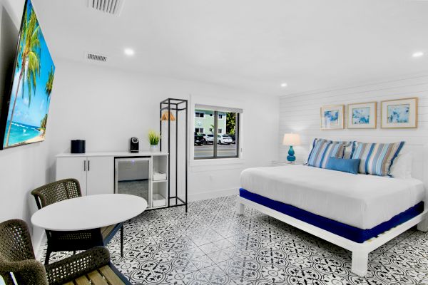 A bright, modern bedroom features a large bed, patterned floor, wall art, a TV, a small dining area, and a window with a view outside.