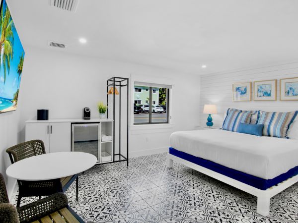 A bright modern hotel room with a white and blue theme: a large bed with blue accents, patterned floor tiles, a small round table with chairs, a wall-mounted TV, and a window showing greenery outside.