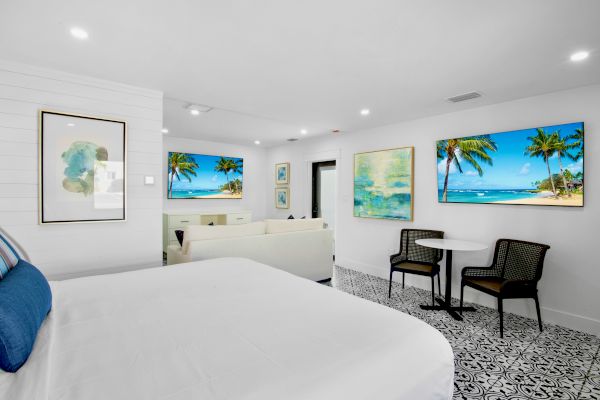 A bright, modern room with a bed, chairs, table, and beach-themed artwork on white walls beneath recessed lighting.