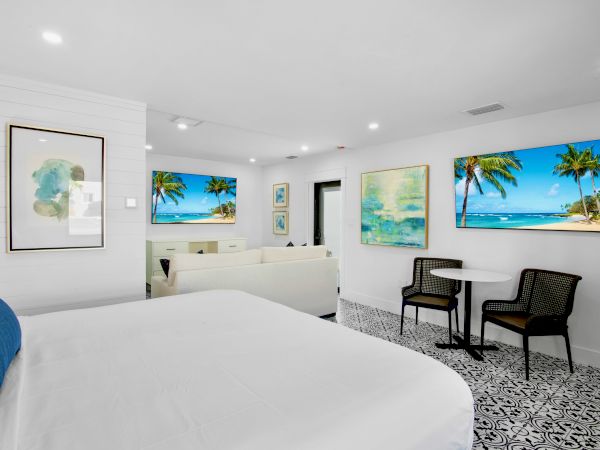 A bright, modern room with a bed, chairs, table, and beach-themed artwork on white walls beneath recessed lighting.