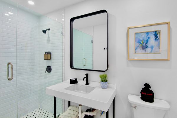 A modern bathroom with a glass shower, sleek sink, potted plant, and artwork above the toilet on a white wall.
