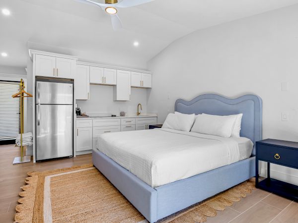 A bright studio with a blue upholstered bed, white linens, a small navy nightstand, a rustic rug, and a compact kitchenette area in the background.