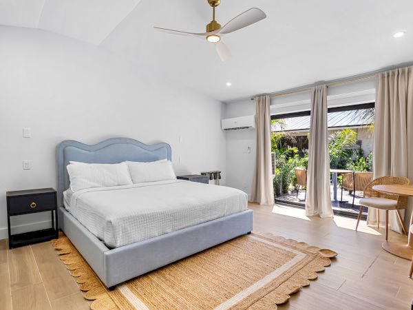 A bright hotel-style bedroom with a pale blue upholstered bed, white bedding, a woven rug, nightstands, a ceiling fan, and large glass doors opening to a balcony.