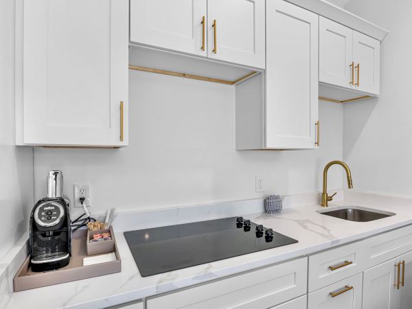 A modern white kitchen with gold hardware, a black cooktop, a sink with a gold faucet, and a coffee station on the counter.