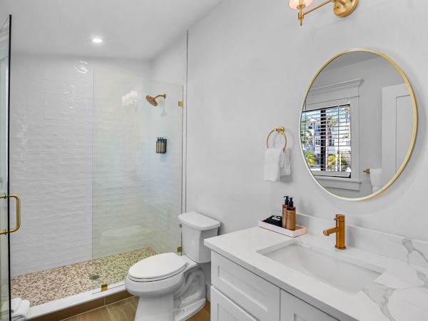 A modern white bathroom with a glass shower, white vanity, round gold-framed mirror, and brass fixtures. Ends with a period.