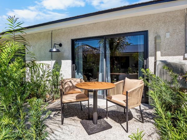 A small outdoor patio with a round wooden table and two beige chairs, surrounded by tall green plants, in front of a sliding glass door.