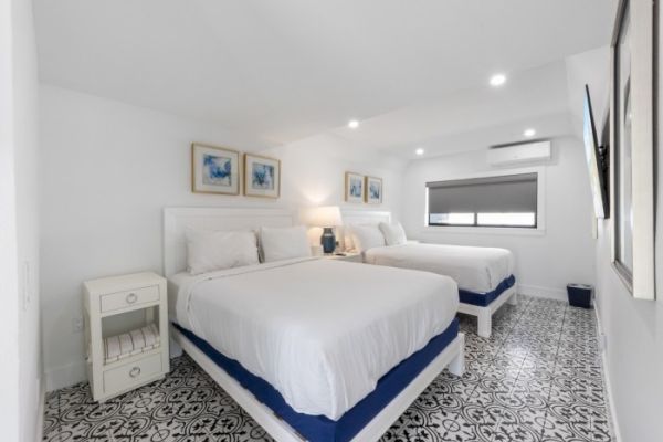 A bright bedroom with two beds, white bedding, wall art, a nightstand, and patterned floor tiles, under soft ceiling lights.