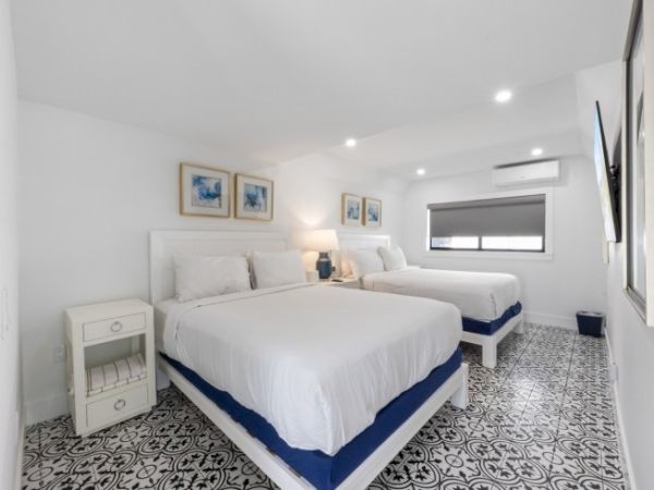 A bright bedroom with two beds, white bedding, wall art, a nightstand, and patterned floor tiles, under soft ceiling lights.
