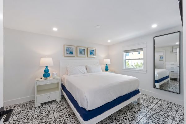 A bright bedroom with patterned floor, double bed, blue lamps, wall art, and large mirror beside a window.