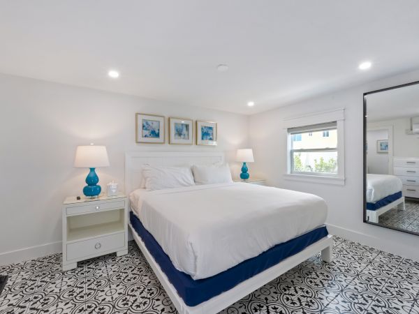A bright bedroom with patterned floor, double bed, blue lamps, wall art, and large mirror beside a window.