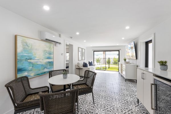 A modern living space with a tiled floor, wicker chairs, a small dining table, wall art, TV, and large windows opening to a garden.