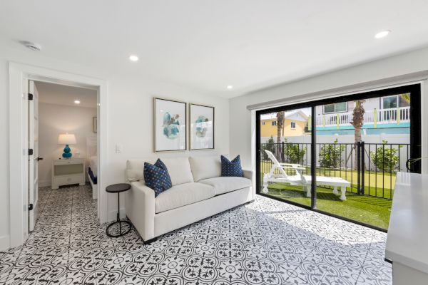 A bright living area with patterned flooring, a white sofa, two blue cushions, and large glass doors opening to a small garden space.