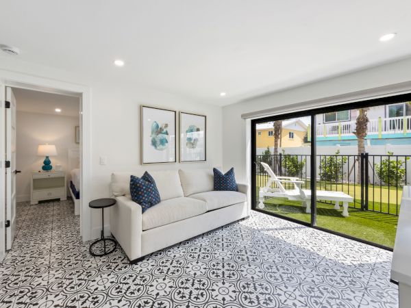 A bright living area with patterned flooring, a white sofa, two blue cushions, and large glass doors opening to a small garden space.