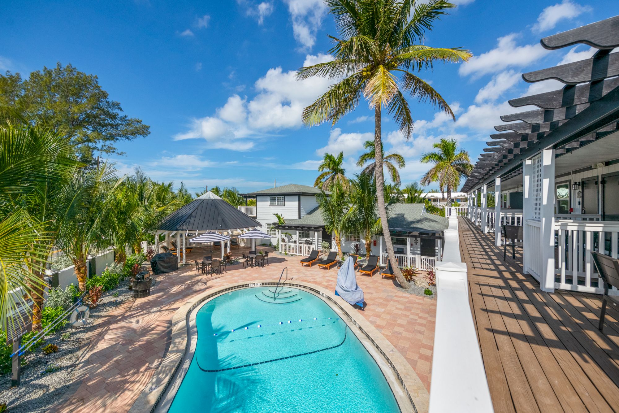 A tropical resort with a swimming pool, surrounded by palm trees and lounge chairs, under a clear blue sky.