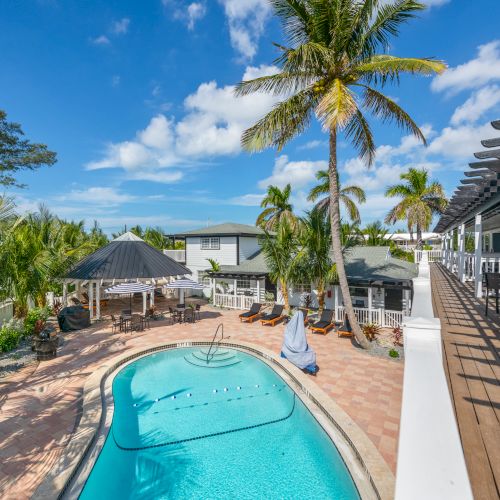 The image shows a tropical outdoor area with a swimming pool, palm trees, a lounge area, and a pergola, under a clear blue sky.