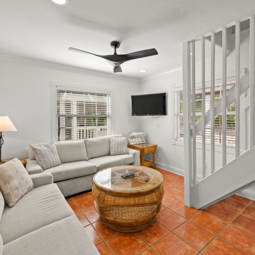 A cozy living room with sofas, a wicker table, lamp, ceiling fan, and TV near stairs, with terracotta tile flooring.