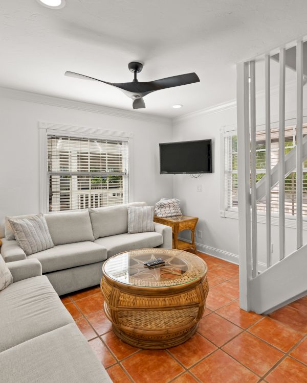 A cozy living room with sofas, a wicker table, lamp, ceiling fan, and TV near stairs, with terracotta tile flooring.