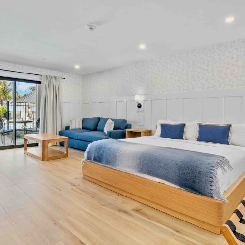 A bright, modern hotel room with a large wooden bed, blue and white bedding, a blue sofa, bedside tables, and a balcony view outside.