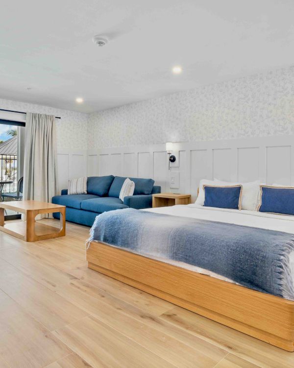 A bright, modern hotel room with a large wooden bed, blue and white bedding, a blue sofa, bedside tables, and a balcony view outside.
