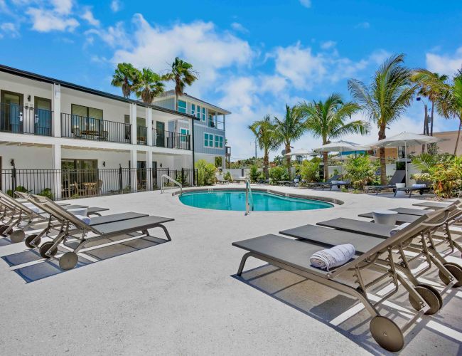 A sunny pool area at a modern hotel with palm trees, lounge chairs, and a round pool, surrounded by a two-story building.