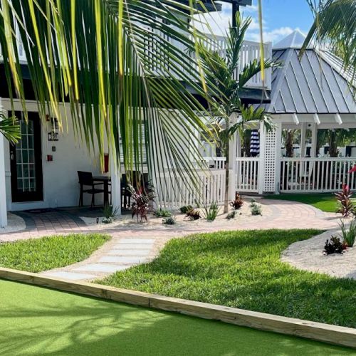 A tropical yard with a mini-golf course, bocce court, and palm trees around a white house.