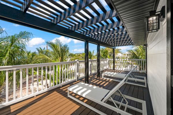 A sunlit patio with two loungers, a white railing, and a pergola, overlooks tropical trees and a clear blue sky.