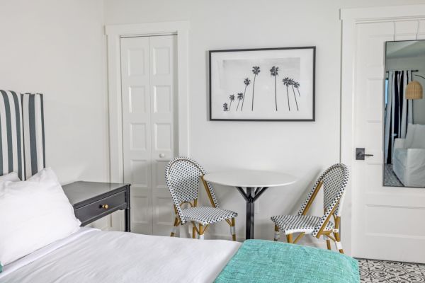 A tidy bedroom with a bed, desk, round table, two chairs, and wall art depicting palm trees.