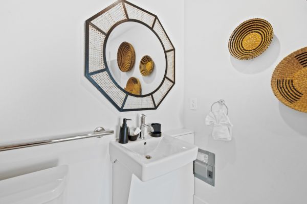 A bathroom with a modern sink, octagonal mirror, wall-mounted woven baskets, soap dispensers, and a toilet handrail is shown.