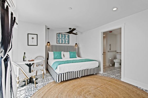 A bright bedroom with a striped bed, teal accents, a small table with chairs, and an adjoining bathroom.