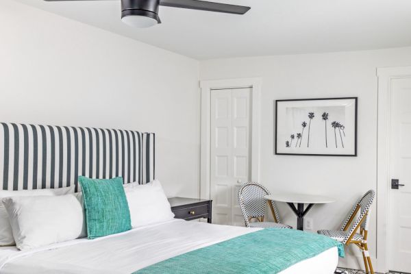 A modern bedroom with a striped headboard, teal accents, wall art, round table, chairs, and ceiling fan, all in a bright, minimalist style.