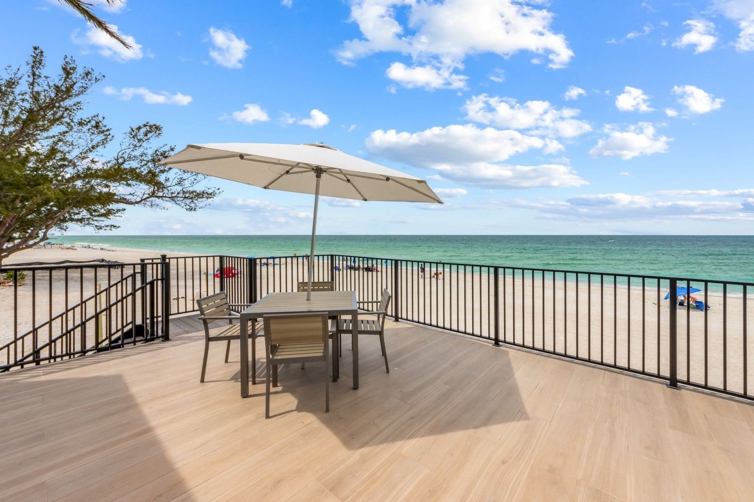 A beachside deck with a table, chairs, and umbrella overlooking a sandy beach and turquoise sea under a partly cloudy blue sky.