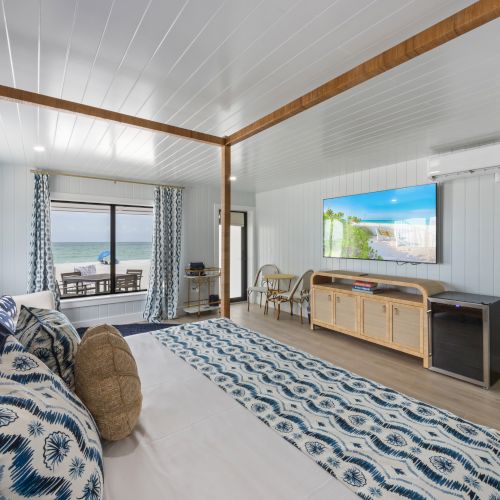 A modern bedroom with ocean view, featuring a bed, TV, blue accents, wooden furniture, and sliding doors leading to a balcony with seating.