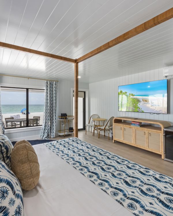 A modern bedroom with ocean view, featuring a bed, TV, blue accents, wooden furniture, and sliding doors leading to a balcony with seating.