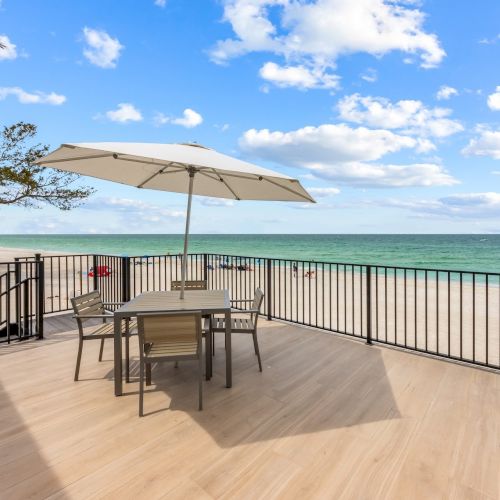 A beach deck with a table, chairs, and umbrella overlooking the ocean. The sky is partly cloudy with trees and beachgoers in the distance.