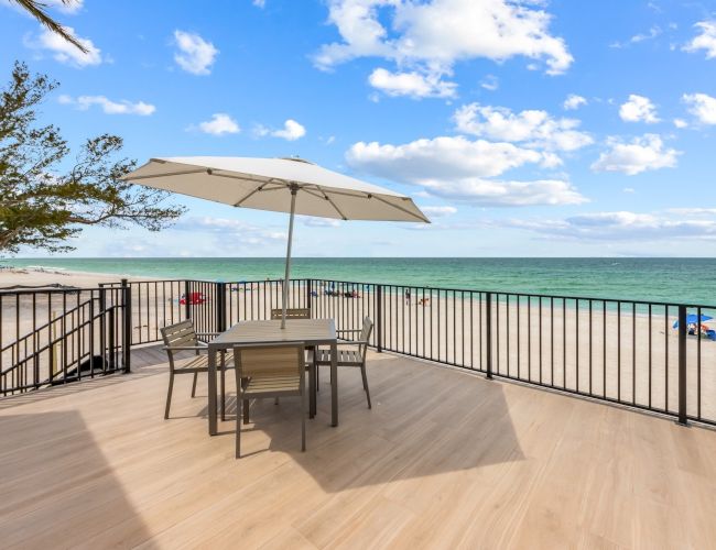 A beach deck with a table, chairs, and umbrella overlooking the ocean. The sky is partly cloudy with trees and beachgoers in the distance.