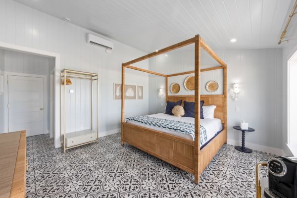 A bright bedroom with a four-poster bed, decorative flooring, and wall art. There's a side table and an open clothing rack.
