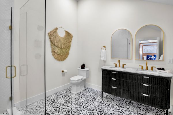 A bathroom with a glass shower, patterned floor tiles, a double vanity with mirrors, and wall decor is shown, featuring golden fixtures.