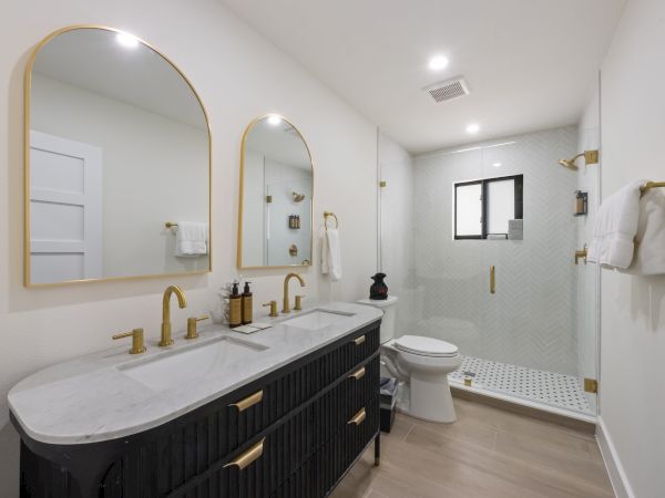A modern bathroom featuring double sinks with gold fixtures, two mirrors, a toilet, and a glass-enclosed shower with elegant decor.