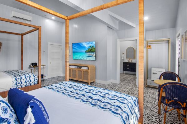 A cozy bedroom with blue accents features a bed, wall-mounted TV, wicker seating, and a tiled floor. There is a visible open doorway.