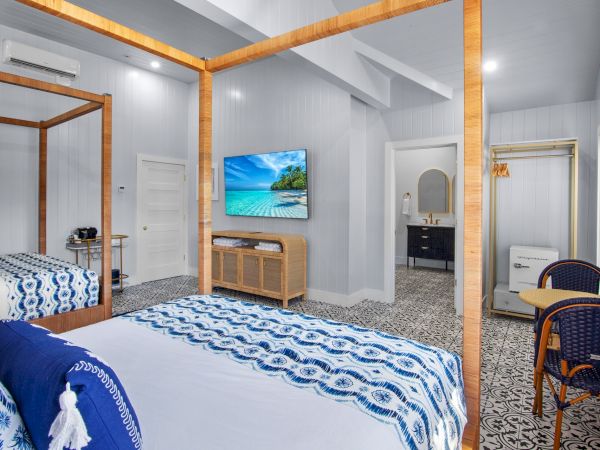 A cozy bedroom with blue accents features a bed, wall-mounted TV, wicker seating, and a tiled floor. There is a visible open doorway.