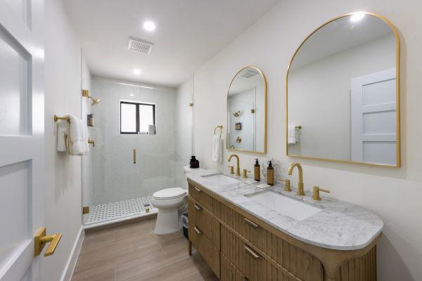 Modern bathroom with double vanity, arched mirrors, gold fixtures, and a walk-in shower. Neutral tones, sleek design, and bright lighting.