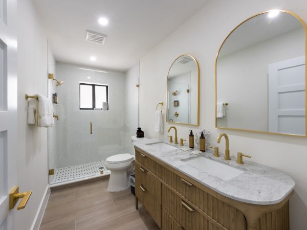 Modern bathroom with double vanity, arched mirrors, gold fixtures, and a walk-in shower. Neutral tones, sleek design, and bright lighting.