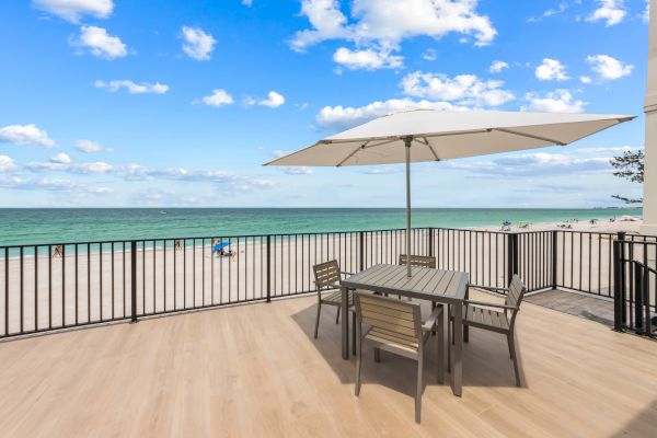 A beach view features a deck with a table, chairs, and an umbrella, overlooking the ocean under a partly cloudy sky.