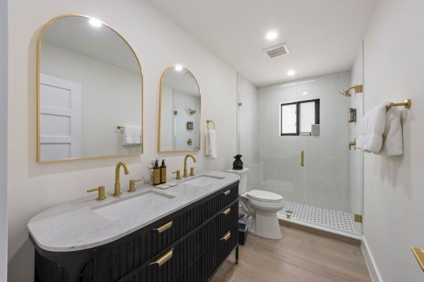 The image shows a modern bathroom with a double vanity, gold fixtures, dual mirrors, a toilet, and a glass-enclosed shower with white tiles.