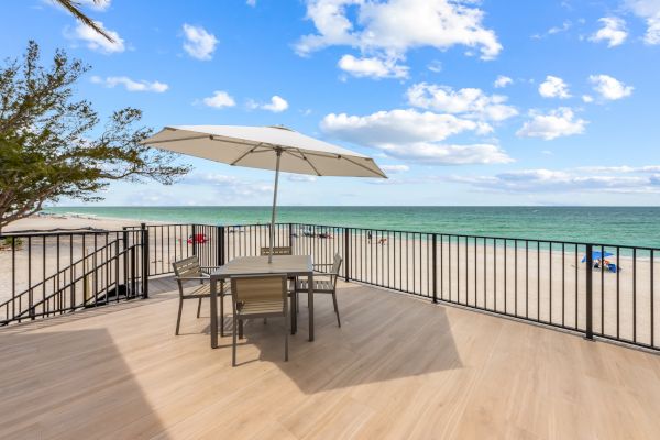 A wooden deck overlooking a beach with a table, chairs, and umbrella under a blue sky with scattered clouds and calm sea.